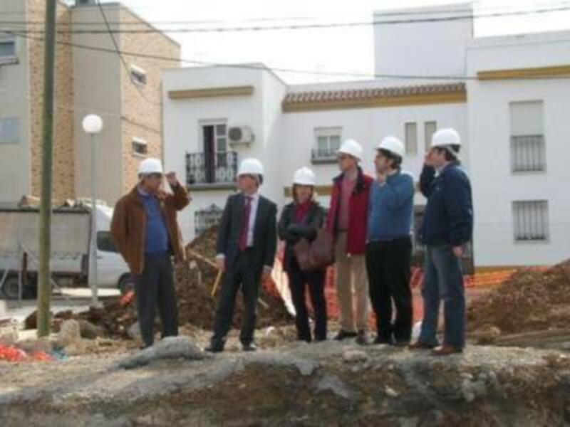 Una grupo de personas con cascos de construcción observan una obra en un entorno urbano. La imagen muestra una calle con edificios y un camión de carga en el fondo, indicando que se está realizando una construcción o reparación.