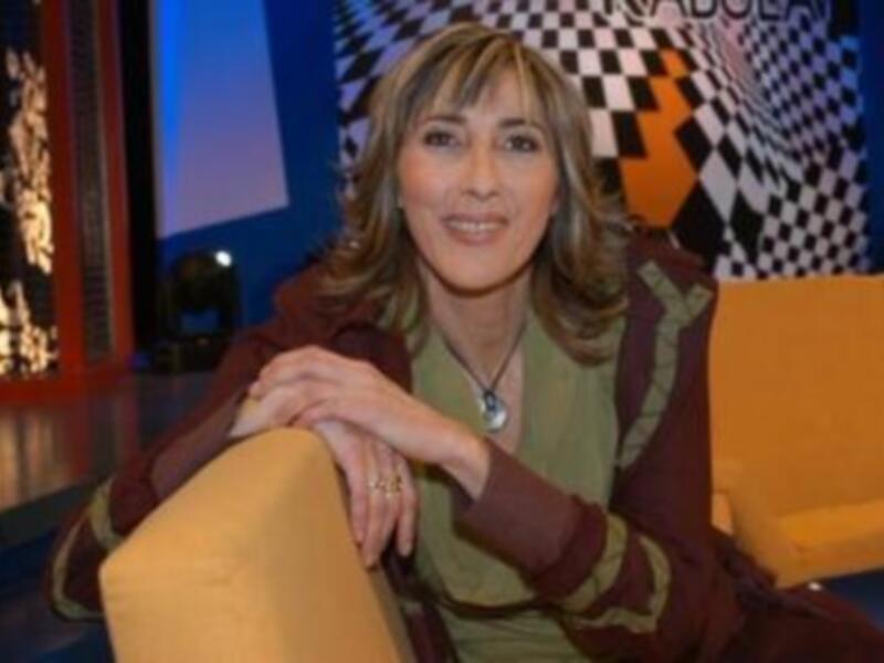 Una mujer con cabello largo y rizado, vistiendo una chaqueta de color marrón y un suéter verde, sentada en un sofá amarillo. La imagen tiene un fondo con patrones geométricos y letras en el techo, probablemente de una televisión o programa.