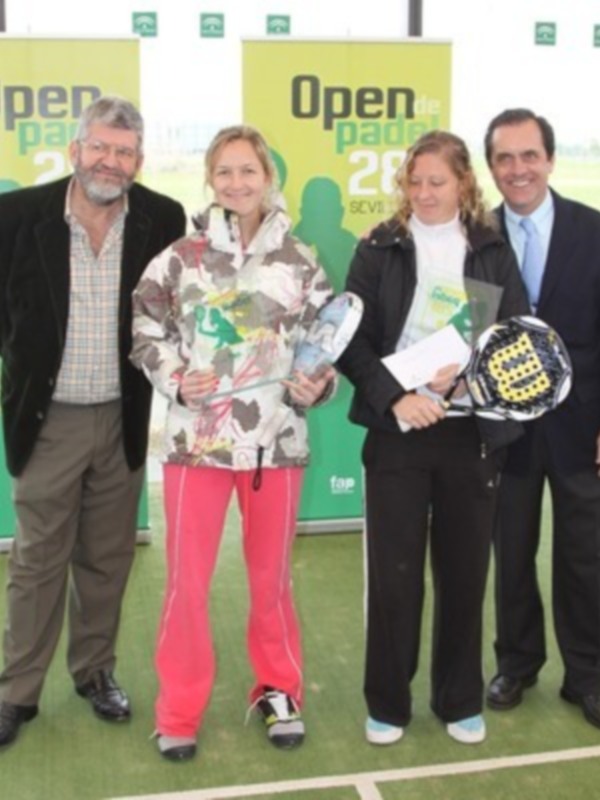 Fotografía de cuatro personas posando en un campo de tenis. En el fondo, dos carteles con la inscripción "Open padel 2018". La persona central sostiene un raqueta de tenis y una placa con un trofeo.