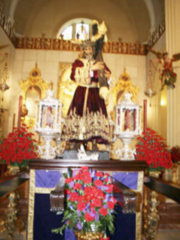 Un altar con una estatua de Cristo en la parte frontal, rodeada por flores rojas y blancas. El altar está decorado con detalles dorados y una araña de cristal en la parte superior. La estatua está vestida con un manto negro y una corona dorada, lo que la hace destacar en el centro del altar. En los laterales, hay dos pequeñas estatuas y una araña de cristal más pequeña. La decoración del altar, junto con la estatua central, sugiere un ambiente religioso y festivo.