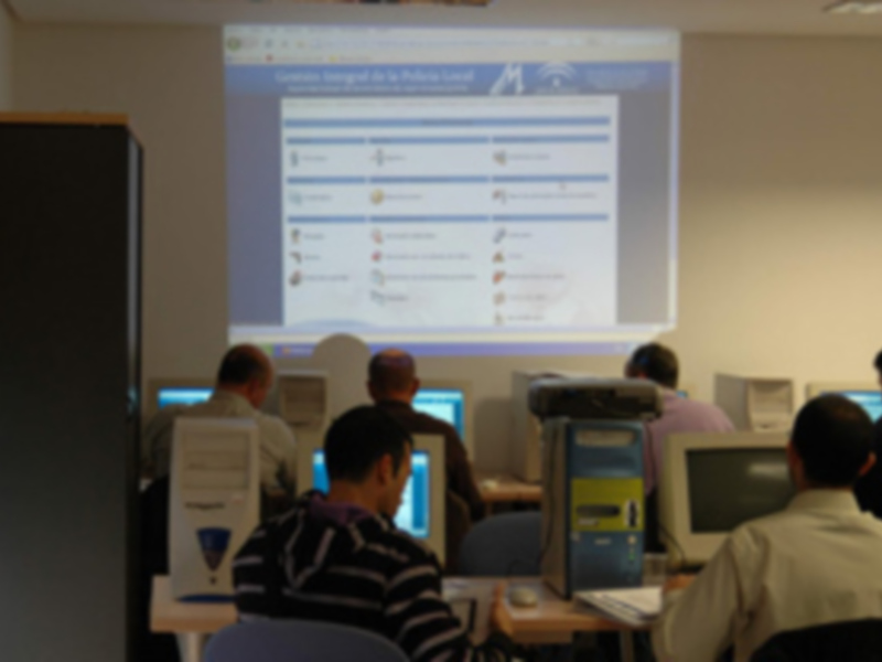 En una sala de computación, varios estudiantes están sentados frente a sus ordenadores, mirando un proyector que muestra una presentación de la "Escuela de Negocios". La atmósfera es de concentración y aprendizaje.