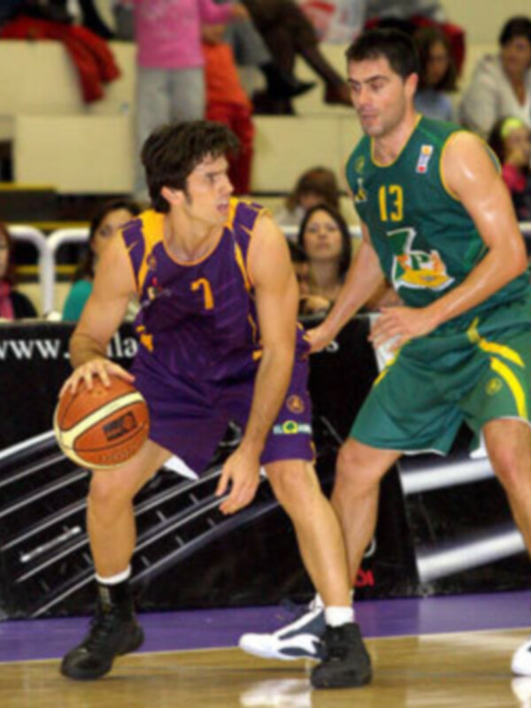Jugadores de baloncesto en acción durante un partido, uno en uniforme violeta y el otro en verde, en un entorno deportivo con público en el fondo.