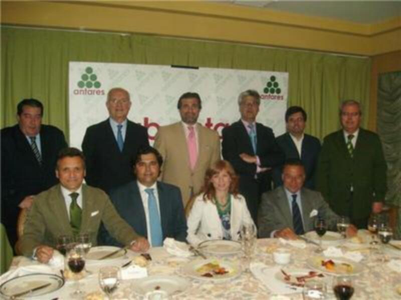 Grupo de personas en una reunión formal, posiblemente un evento empresarial o de networking. En el fondo, hay una pantalla con la marca "antares" y un logo triangular verde claro. La mesa está servida con copas de vino, platos y servilletas.