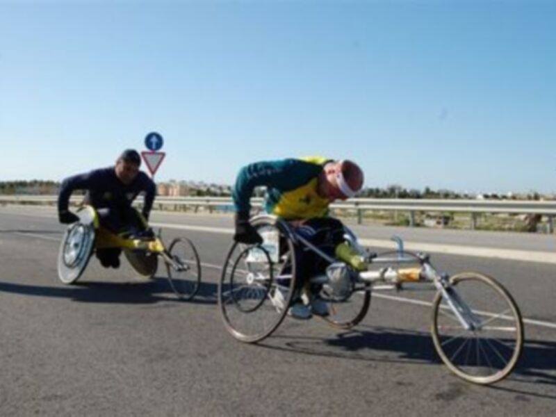 Dos ciclistas en sillas de ruedas competiendo en una carretera de asfalto, con un cielo azul claro y un camino de tierra en el fondo.