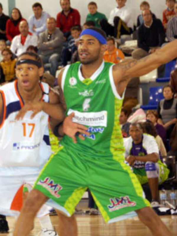 Un jugador de baloncesto en acción durante un partido, con otro rival intentando bloquear su tiro. El jugador del equipo verde lleva una camiseta con el nombre 'Schachmied' y los números 17 y 24, mientras que el rival lleva una camiseta con 'JIM'.