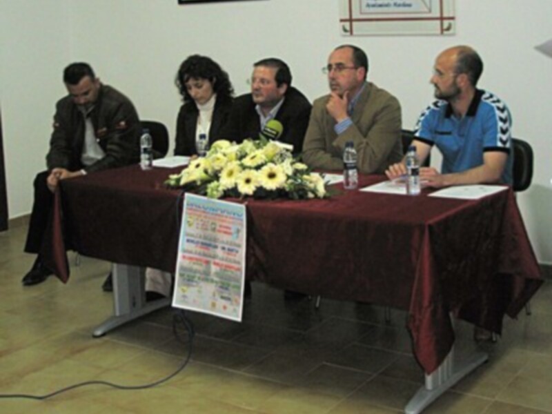 Un grupo de personas se sientan en una mesa con un mantel rojo, rodeada por flores y un cartel informativo.
