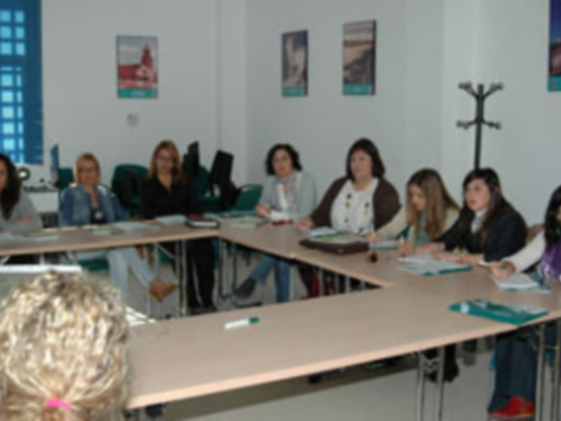 Una clase de idiomas con estudiantes sentados en mesas, un profesor frente a ellos. La sala tiene ventanas azules y carteles en las paredes.