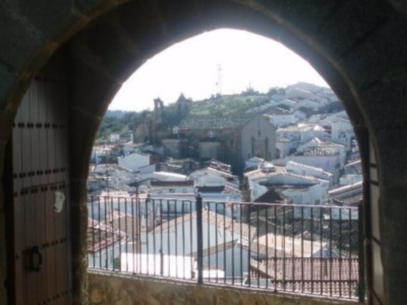 Vista desde una ventana de una fortaleza hacia una ciudad con edificios blancos y verdes montañas en el fondo.