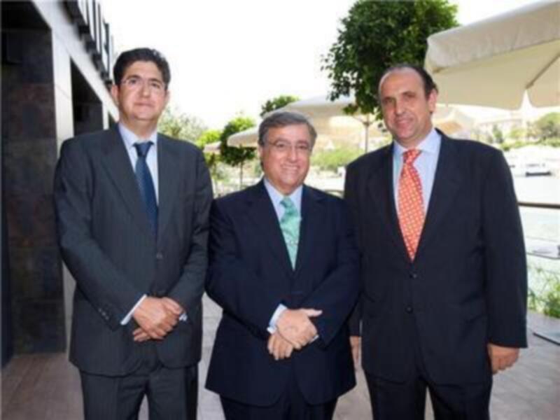 Tres hombres en trajes posan juntos, uno de ellos estrecha la mano de uno de los otros dos, en un ambiente formal con una sombrilla en el fondo.