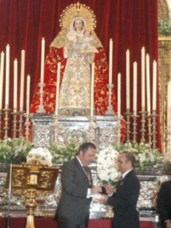 Dos hombres en trajes se encuentran frente a una imagen religiosa, rodeados de flores y velas.