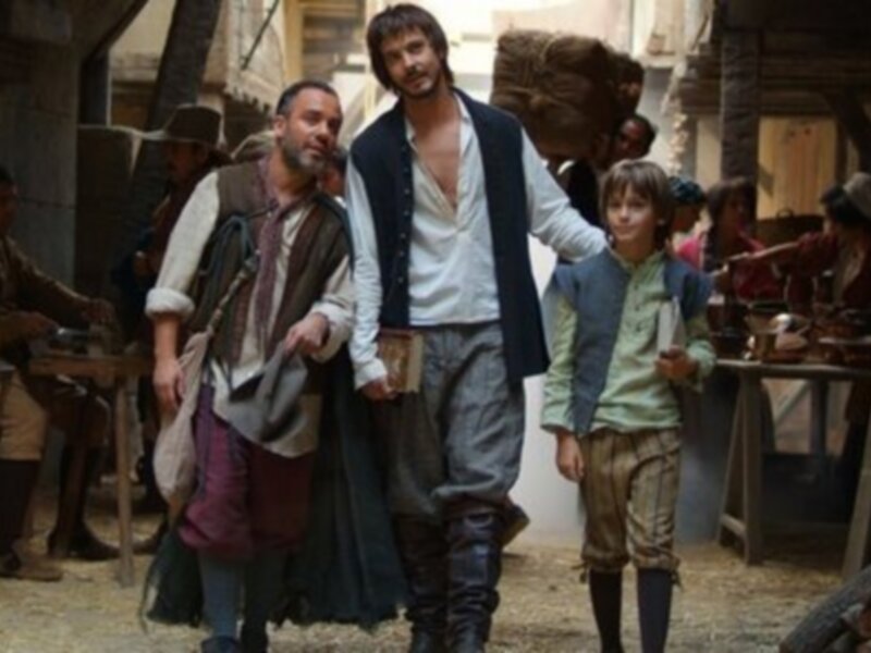Imagen de tres personajes en traje histórico, posiblemente de época medieval. Dos hombres y un niño. Los hombres llevan vestimentas con detalles de cuero, mientras que el niño usa pantalones cortos y una chaqueta azul. La escena parece ser en un entorno de mercado o calle con estructuras de madera y tiendas.