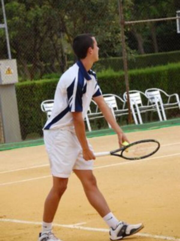 Un joven vestido con una camiseta azul y blanca, pantalones cortos blancos y zapatillas de deporte blanco y negro está en una cancha de tenis, preparándose para un golpe.