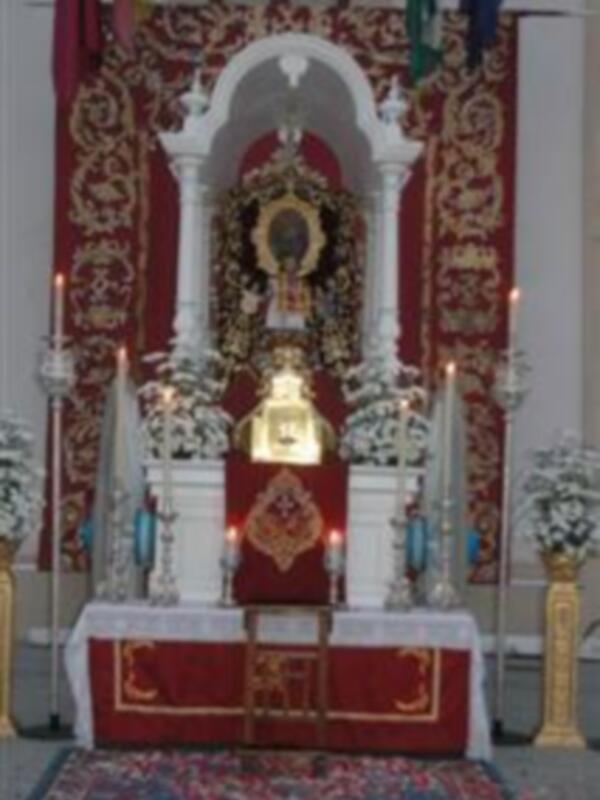 Un altar con una imagen de la Virgen en el centro, rodeado por flores y velas encendidas. La decoración incluye banderas y mantones de color rojo y dorado, creando un ambiente religioso y festivo.