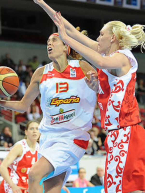 Dos jugadoras de baloncesto en acción, una con el número 13 y la marca "España" en su camiseta, intentando bloquear a otra jugadora con el número 1. El fondo muestra una arena deportiva llena de espectadores.