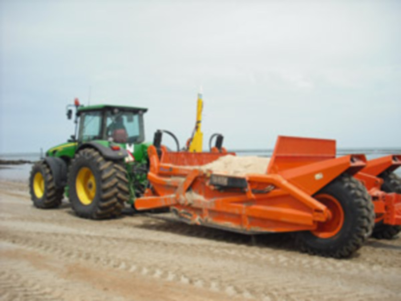 Un tractor agrícola con un remolque para transporte de materiales en una playa arenosa, ilustrando el uso de maquinaria agrícola en áreas no convencionales.