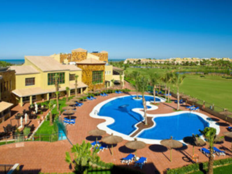 Un resort con piscina en forma de corazón, rodeada por palmeras y sombrillas. La vista es maravillosa con un campo de golf en primer plano y una costa al fondo.