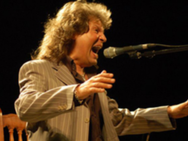 Un hombre con cabello largo y barba, vestido de gris, canta en un escenario con un micrófono en frente.