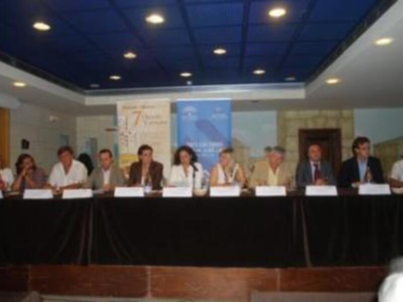 Una conferencia de prensa con varios presentadores sentados en una mesa, con un fondo que incluye un cartel con el texto '17º Aniversario' y una imagen de un edificio.