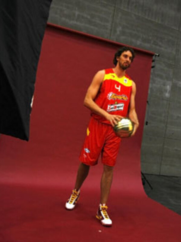 Un jugador de baloncesto en uniforme rojo y amarillo sostiene un balón, posando frente a una pantalla de fondo roja en un estudio fotográfico.