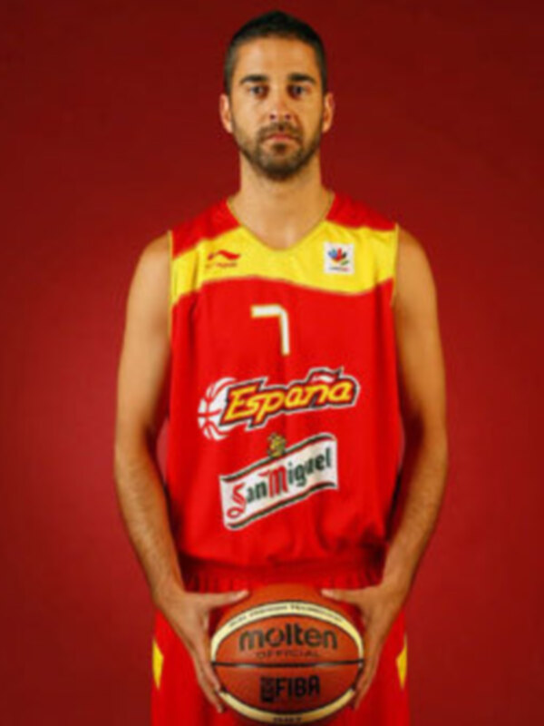 Un jugador de baloncesto posa con una camiseta roja y amarilla que dice "España" en el centro, con un logo de San Miguel en la parte inferior. Lleva una camiseta roja y amarilla con el número 7, sosteniendo un balón de baloncesto. El fondo es rojo oscuro.
