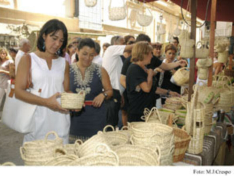 Una joven en un vestido blanco camina entre cestas de mimbre, mientras otras personas observan y conversan en un mercado al aire libre. La imagen captura la vida cotidiana de una feria o tianguis, con productos naturales y artesanías en el centro.