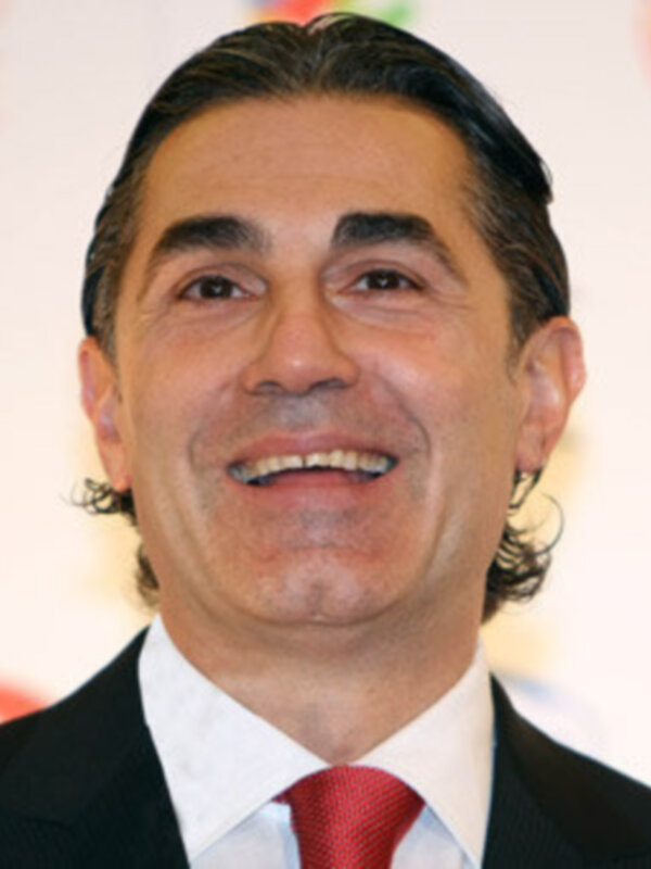 Un hombre con cabello oscuro y largo, sonriendo ampliamente. Lleva una camisa blanca con un cuello de pico y una corbata roja. El fondo es blanco, con un logo en la parte superior derecha.