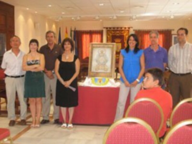 Una reunión en un salón con una mesa redonda, decorada con un pastel y un cuadro. Personas de diferentes edades y estilos vestidos, algunas sonrientes, otras en silencio. En el fondo, cortinas y una bandera.