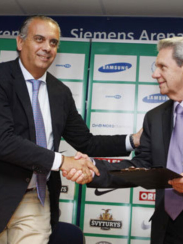 Dos hombres en trajes intercambian un apretón de manos y un documento en un evento en la Siemens Arena.