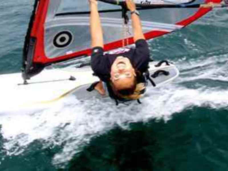 Una persona realiza un salto en una tabla de windsurf, con el viento y la onda del mar detrás.