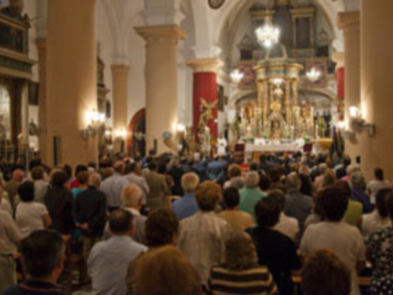 Interior de una iglesia con una gran imagen de la Virgen en el altar, rodeada de fieles en oración. Las columnas y el techo son de estilo barroco, con detalles dorados y luces que iluminan el ambiente.