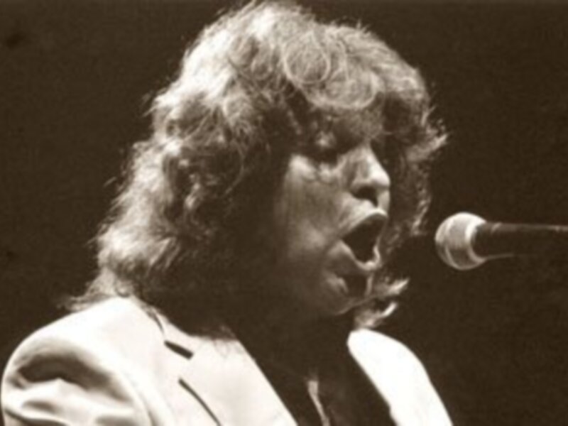Un hombre con cabello largo y una camisa blanca, cantando en un escenario con un micrófono en frente.