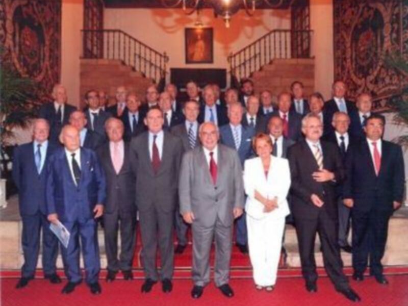 Grupo de personas en trajes formales posando para una foto en un entorno formal, con elementos decorativos y una escalera en el fondo.