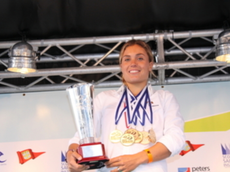 Una mujer sonríe mientras sostiene un trofeo grande y brillante. Detrás de ella, hay una pared con banderas y un logotipo de Peters.