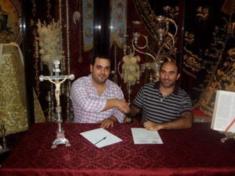 Dos personas firmando un documento en una mesa con un fondo festivo, posiblemente en una ceremonia religiosa o formal.