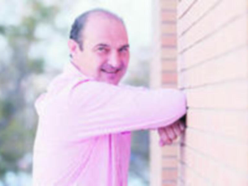 Un hombre en una camisa rosa está mirando por la ventana de un edificio. La imagen parece ser de una escena urbana o residencial, con detalles como la mampara de la ventana y el fondo borroso.