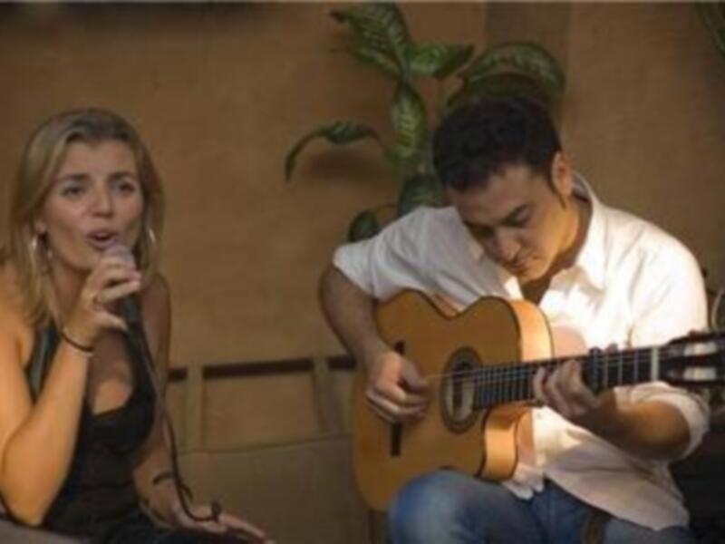 Una joven canta mientras un hombre toca la guitarra acústica. Ambos están sentados frente a una pared de fondo con un gran plátano verde en la esquina.