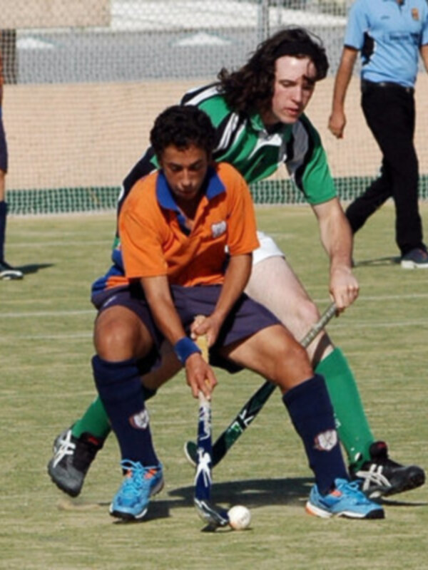 Jugadores de hockey en acción sobre un campo verde, con una figura femenina en el centro y otro jugador detrás.