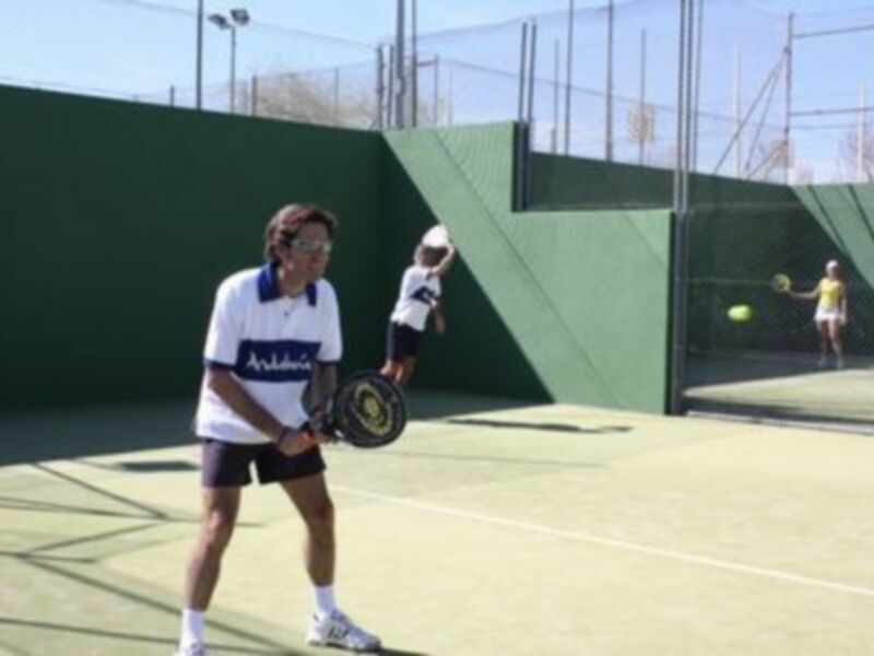 Un hombre con una camiseta blanca y un niño en el fondo juegan al tenis. El hombre está preparado para recibir la pelota, mientras que el niño parece estar en acción de lanzarla. La cancha es verde y el cielo está claro, lo que sugiere un día soleado.