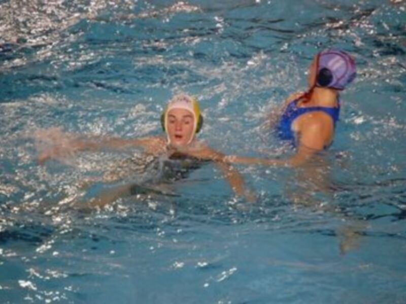 Dos nadadoras en un estanque de agua azul, una con gorro de natación amarillo y la otra con gorro de natación azul, nadan en dirección opuesta.