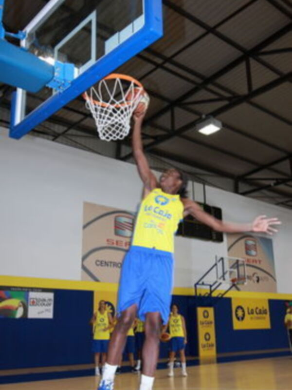 Un jugador de baloncesto en acción, lanzando el balón hacia el aro. El jugador lleva una camiseta amarilla con el nombre del equipo 'EC Club' y pantalones azules. El fondo muestra una cancha de baloncesto con banners de diferentes empresas.