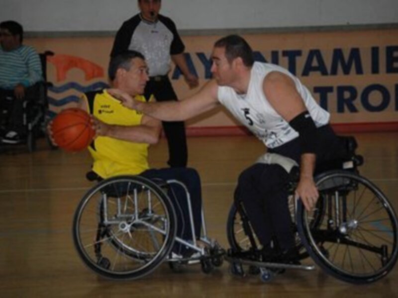 Dos hombres en sillas de ruedas jugando baloncesto, uno con un balón y otro con una camiseta amarilla.