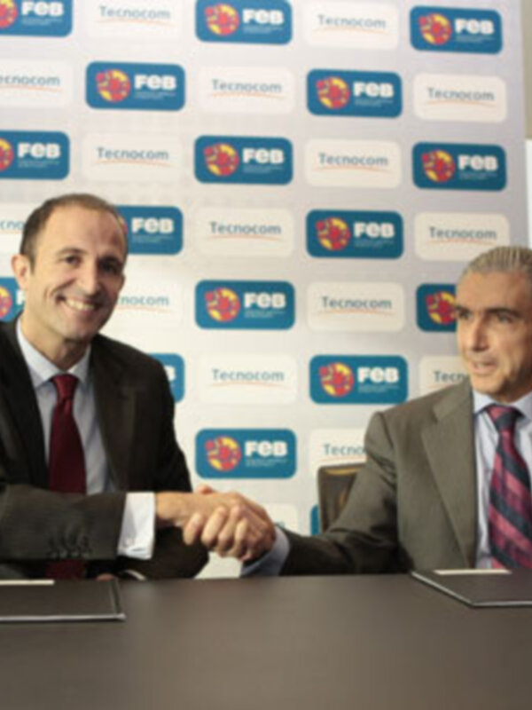 Dos hombres en trajes sentados a una mesa, uno sonriendo y el otro con expresión seria. El fondo es un papelón con logos de FEB Tecnocom repetidos.