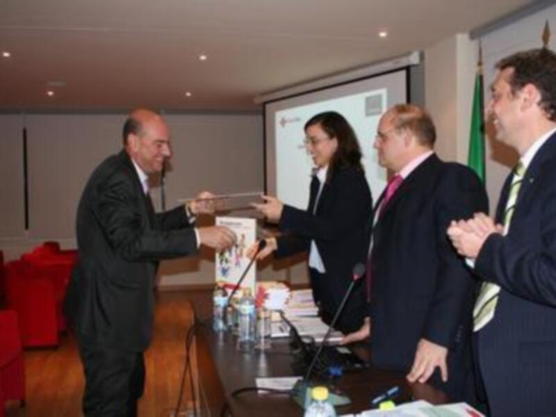 Presentación en conferencia, hombre entrega un libro a una mujer, otros dos hombres observan.