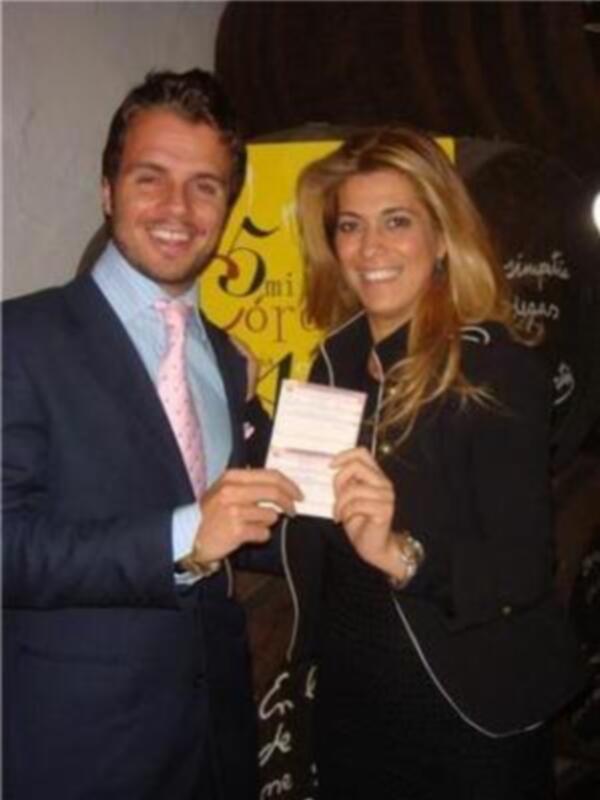 Una pareja sonriente, posando con un documento en la mano, frente a un fondo con letras grandes y un logo.
