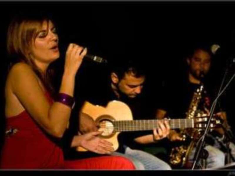 Una mujer en un vestido rojo canta al micrófono mientras un hombre toca la guitarra y otro toca el saxofón en una escena de concierto.