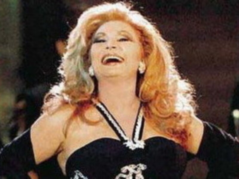 Una mujer con cabello rojo largo y ondulado, vestida con un vestido negro de corte atemporal, posa con una expresión radiante y una sonrisa amplia. La imagen sugiere un ambiente festivo o ceremonial, con luces y sombras que destacan su figura.
