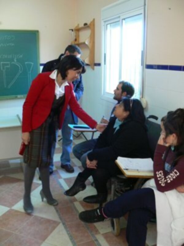 En una aula con pizarra verde, un estudiante en rojo habla con otro sentado en una silla. La escena muestra un ambiente de aprendizaje activo y colaborativo entre estudiantes.