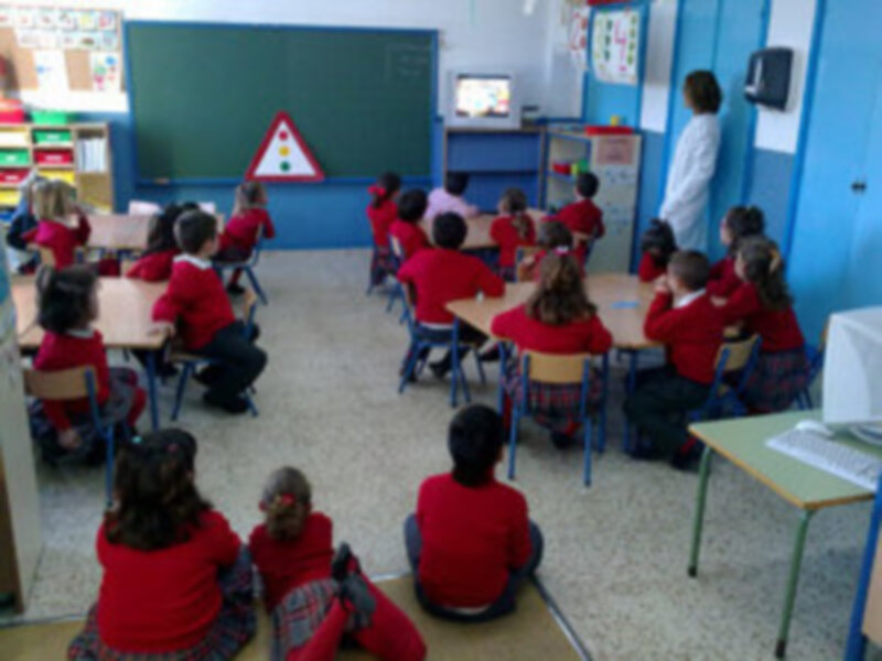 Niños en una clase de educación infantil, sentados en sus respectivos escritorios. En la pared hay un cartel con una señal de tráfico y una imagen en el televisor.