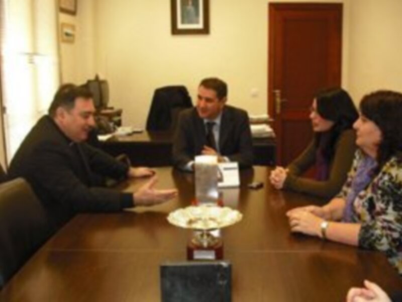Una reunión formal en una oficina con cuatro personas sentadas alrededor de una mesa. La imagen muestra a dos hombres y dos mujeres en traje, discutiendo algo mientras toman notas. La oficina tiene una silla de conferencia y un escritorio con documentos.