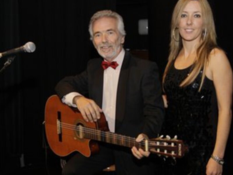 Un hombre en traje y corbata roja toca una guitarra mientras una mujer joven con vestido negro lo mira sonriendo. Ambos están en un escenario con una iluminación suave y fondo oscuro.